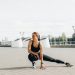 Young woman in black sportswear stretching outdoors in a sunny urban environment.