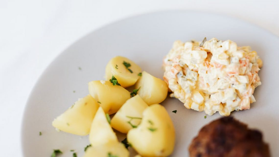 From above of plate with juicy fried meat cutlet served with delicious Russian meat salad and boiled potatoes topped with parsley placed on white table